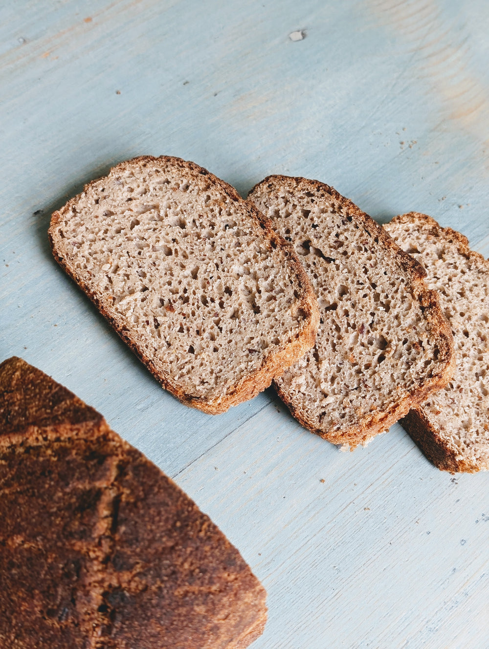 Three slices of whole-grain bread and a partial loaf on a light blue wooden board