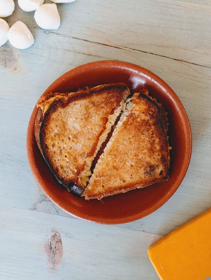 Halved grilled-cheese on brown ceramic plate with melted cheese between halves; mozzarella balls and cheddar wedge at edges.