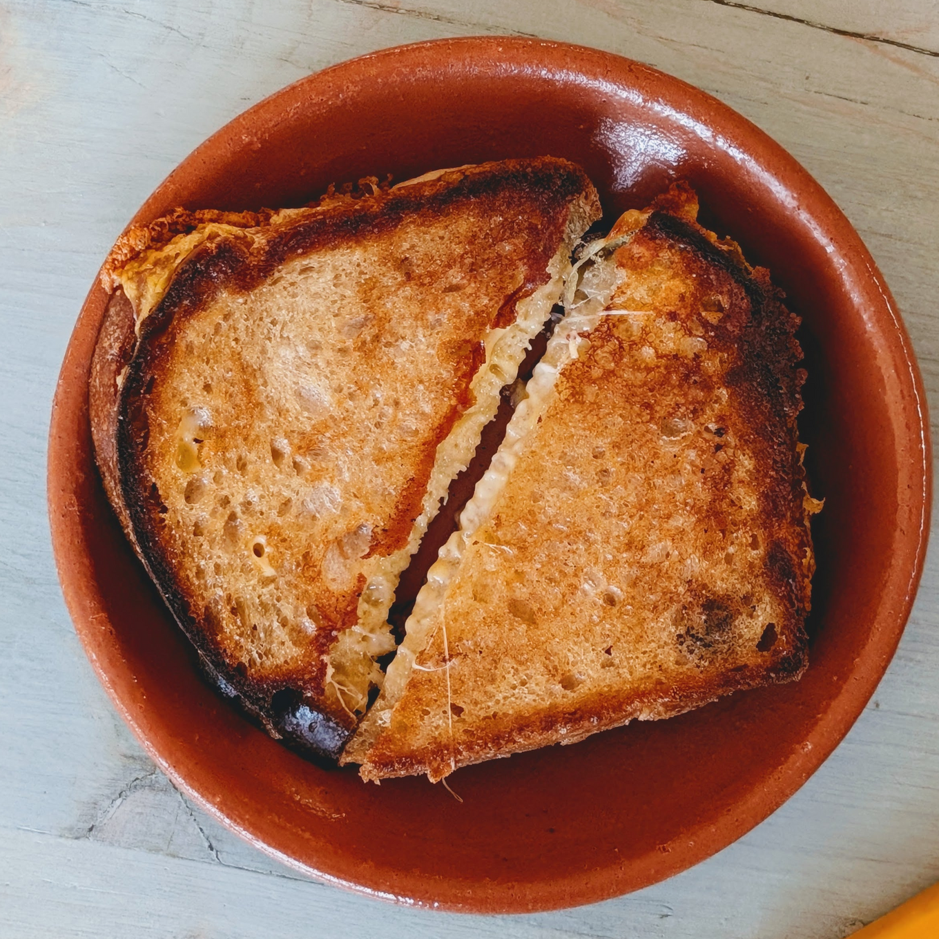 Toasted grilled cheese sandwich cut in half, melted cheese visible, served in a round terracotta bowl on light wood.