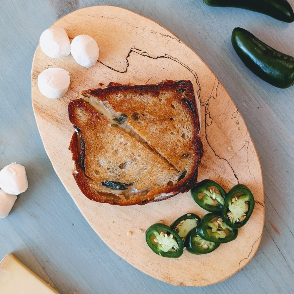 Grilled cheese sandwich, halved and toasted, on a wooden serving board with sliced jalapeños and three small rounds of cheese