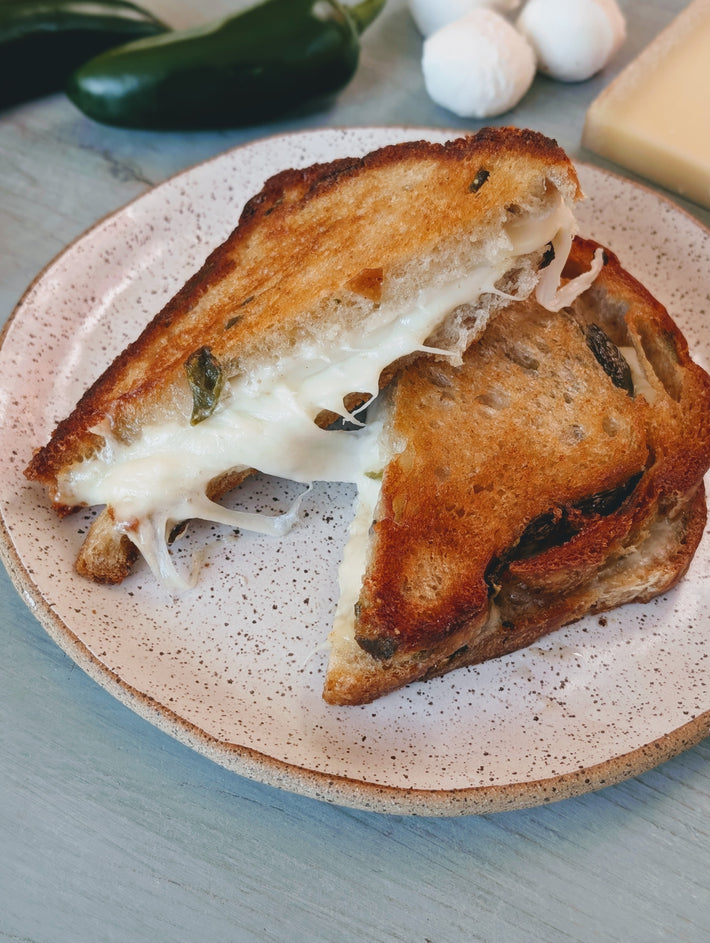 Toasted grilled cheese on herbed sourdough with melted cheese stretching between slices, served on a speckled ceramic plate.