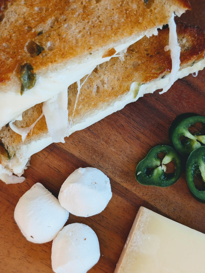 Grilled cheese with gooey melted cheese, three small mozzarella balls, sliced jalapeño rounds and cheese on a wooden board.