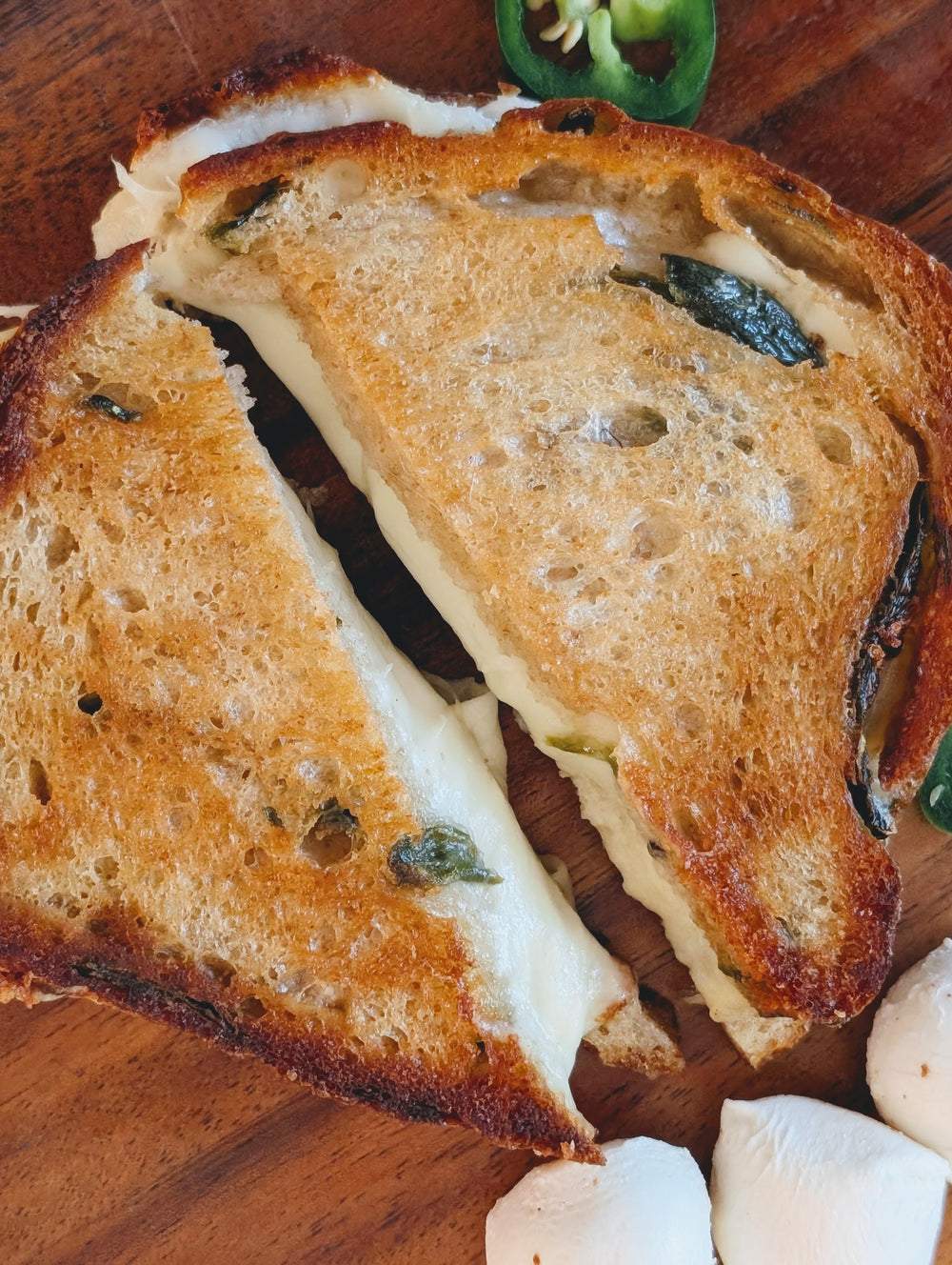 Grilled cheese sandwich with melted cheese and jalapeño, halved on a wooden board next to small mozzarella balls.