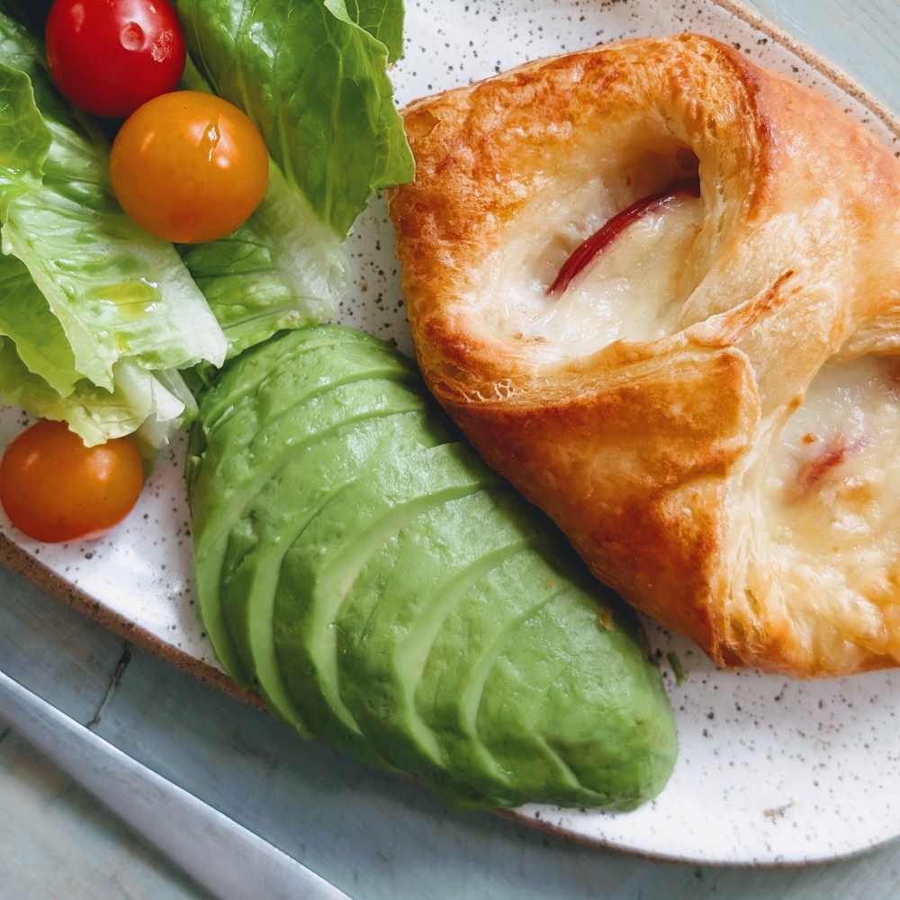 Sliced avocado half, cheese-filled pastry, and lettuce with cherry tomatoes on a speckled plate