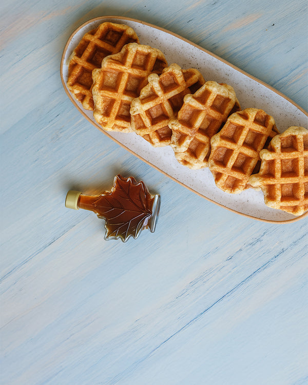 Six golden mini waffles on an oval plate with a maple-leaf syrup bottle beside them on a light blue wooden surface.