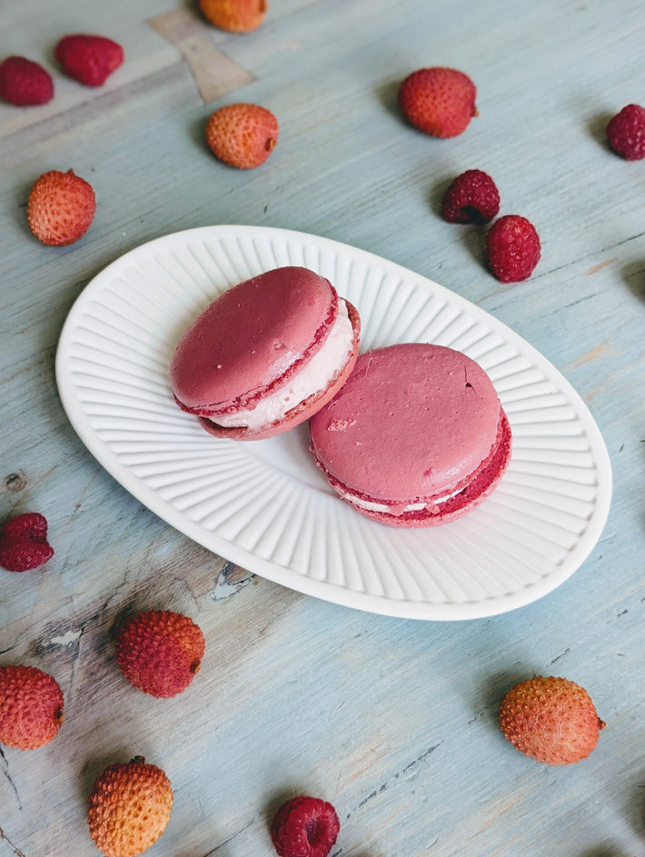 Two pink macarons on a white oval plate surrounded by scattered raspberries and lychees on a blue wooden surface.