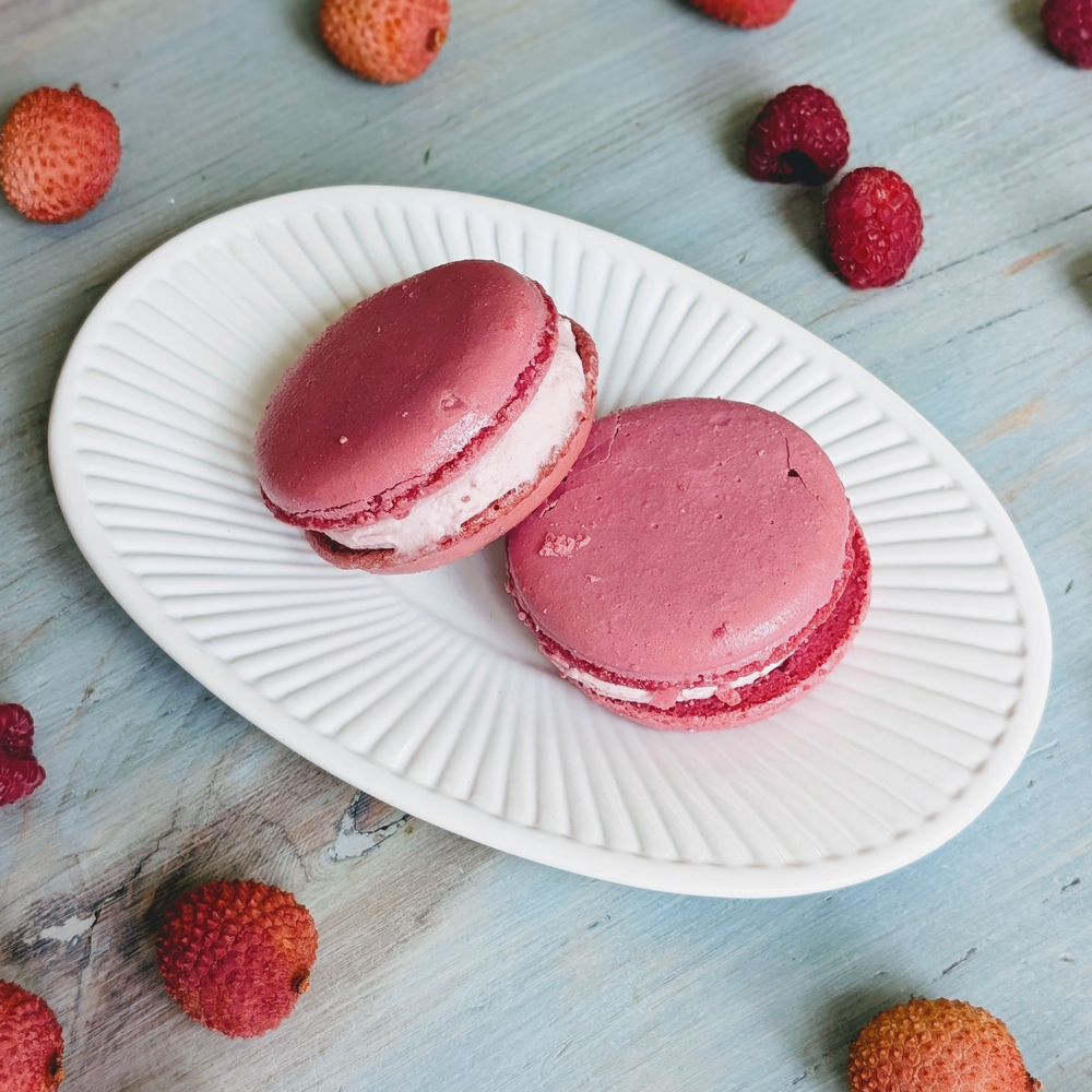 Two pink raspberry macarons with white filling on a fluted white oval plate, raspberries and lychees on blue wooden table.