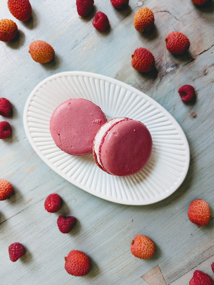Two pink macarons on a white plate surrounded by fresh raspberries and strawberries on a light wooden surface.