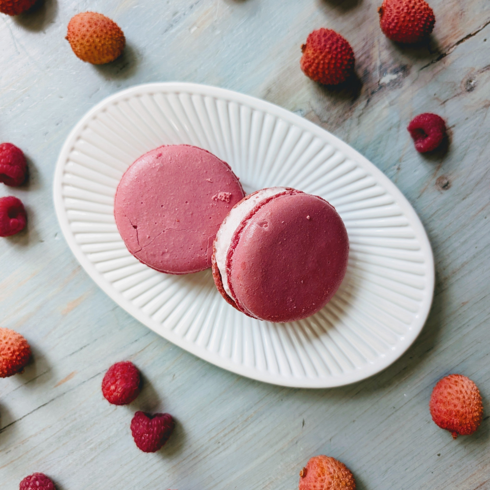 Two pink raspberry macarons on a ridged white oval plate, surrounded by scattered raspberries and lychees on blue wood