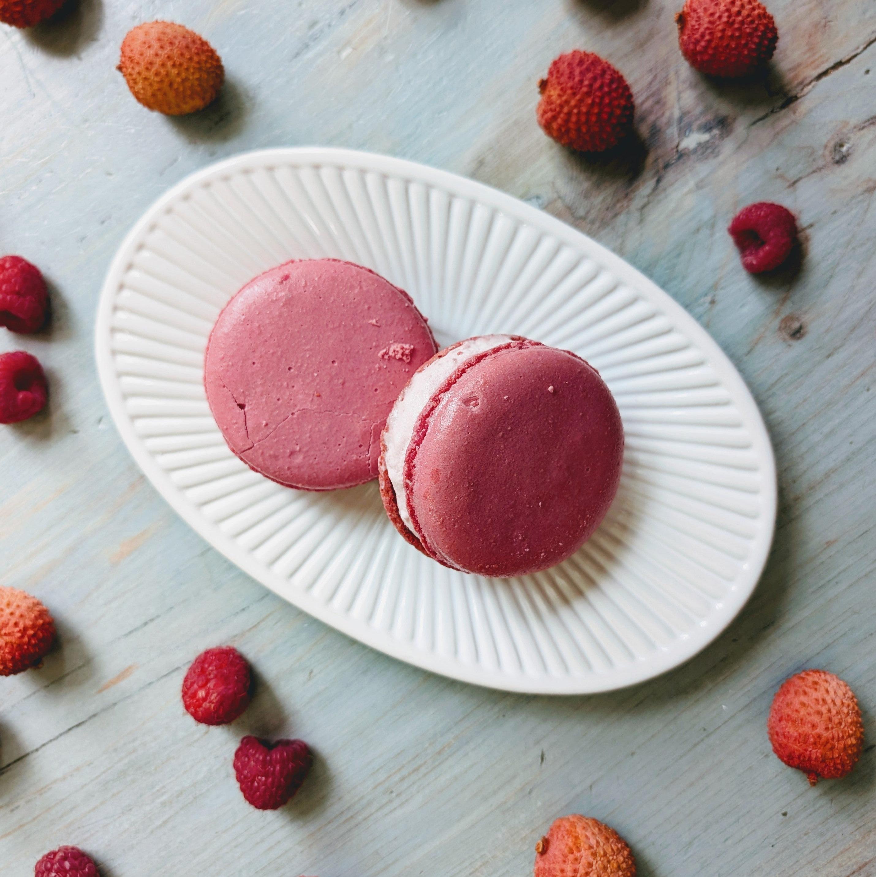 Two pink raspberry macarons on a ridged white oval plate, surrounded by scattered raspberries and lychees on blue wood