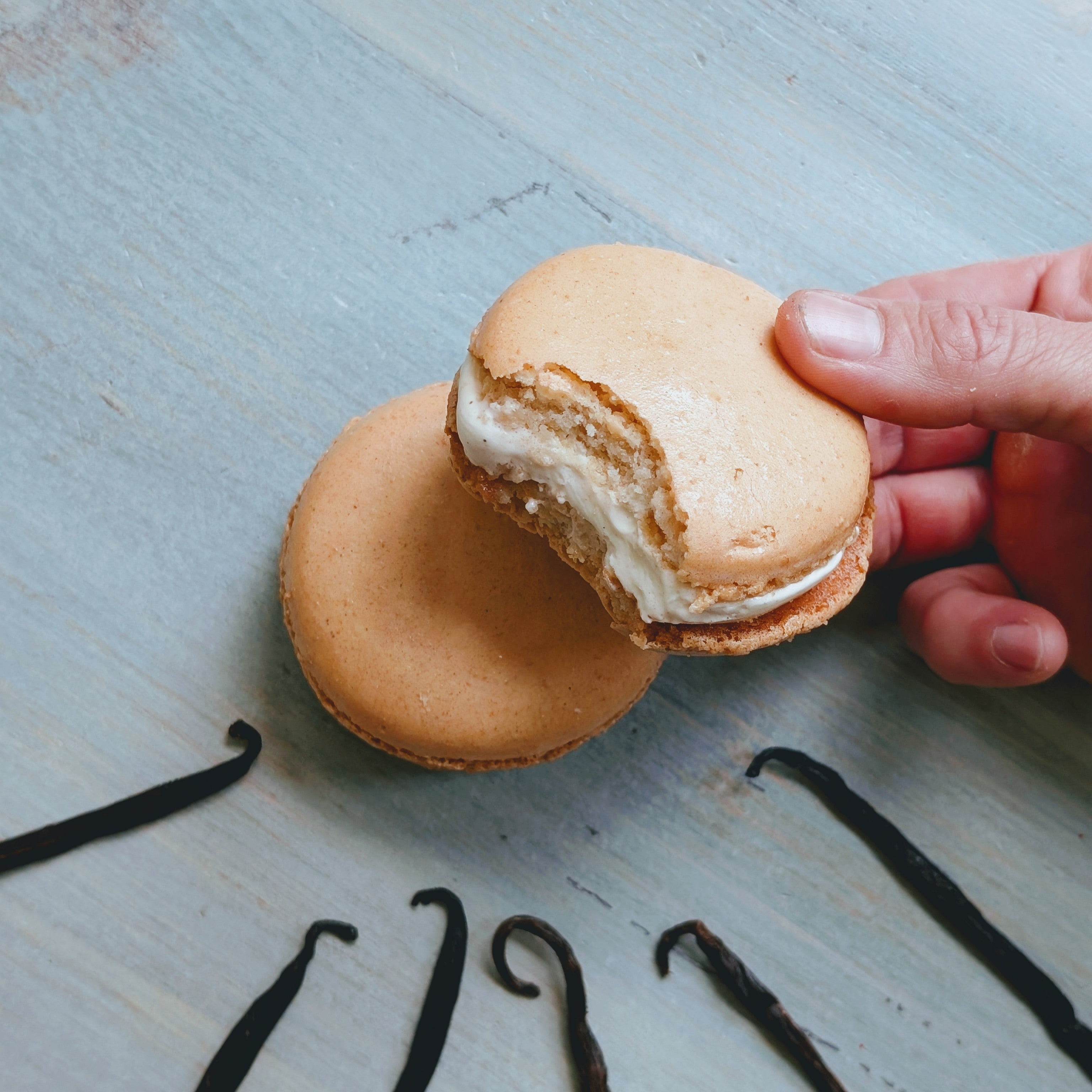 Two vanilla macarons on pale blue wood; one bitten and held by a hand, creamy filling visible; vanilla beans arranged below.