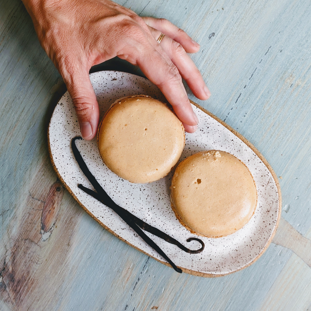 Hand reaching for two beige macarons on a speckled oval plate with two vanilla beans on a pale blue wooden table.