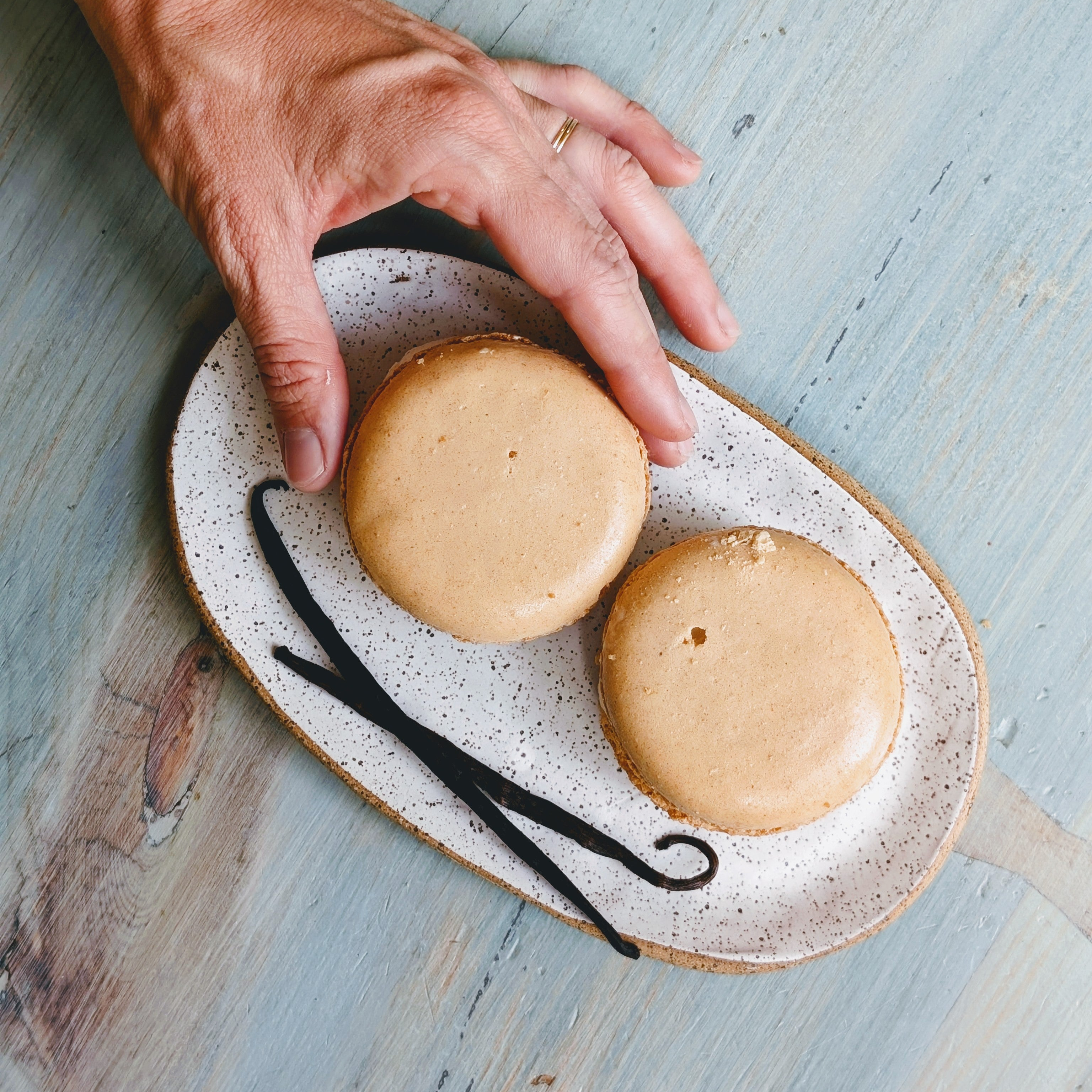 Hand reaching for two beige macarons on a speckled oval plate with two vanilla beans on a pale blue wooden table.