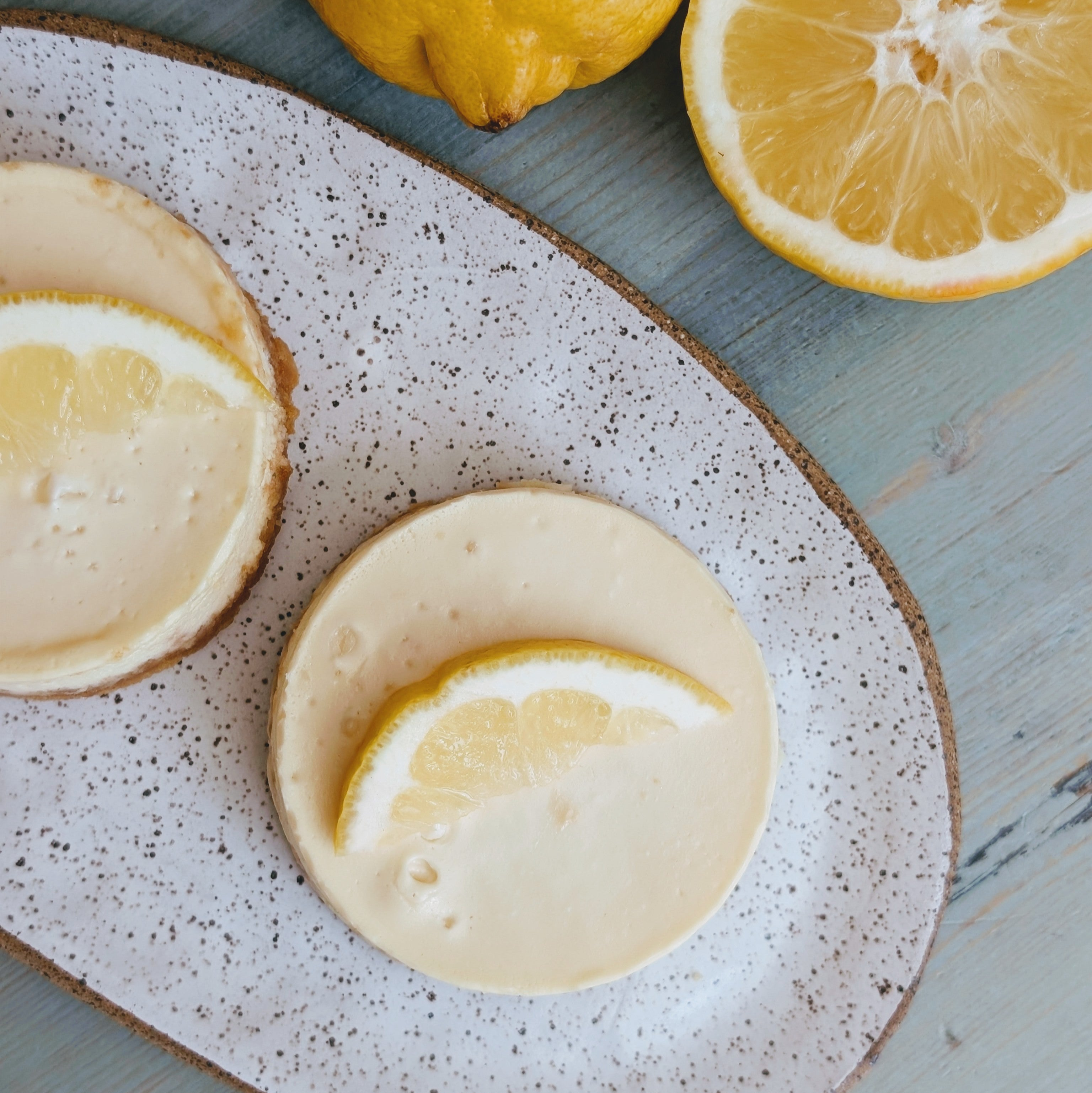 Two round lemon tarts topped with lemon slices on a speckled ceramic platter, with halved lemons on a wooden table.