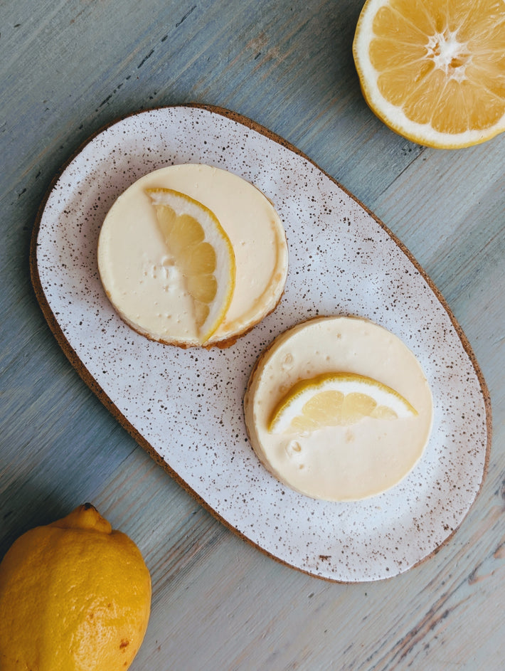 Two lemon slices on top of two round Wildgrain Lemon Cheesecake on a speckled ceramic plate with lemons on a wooden surface.