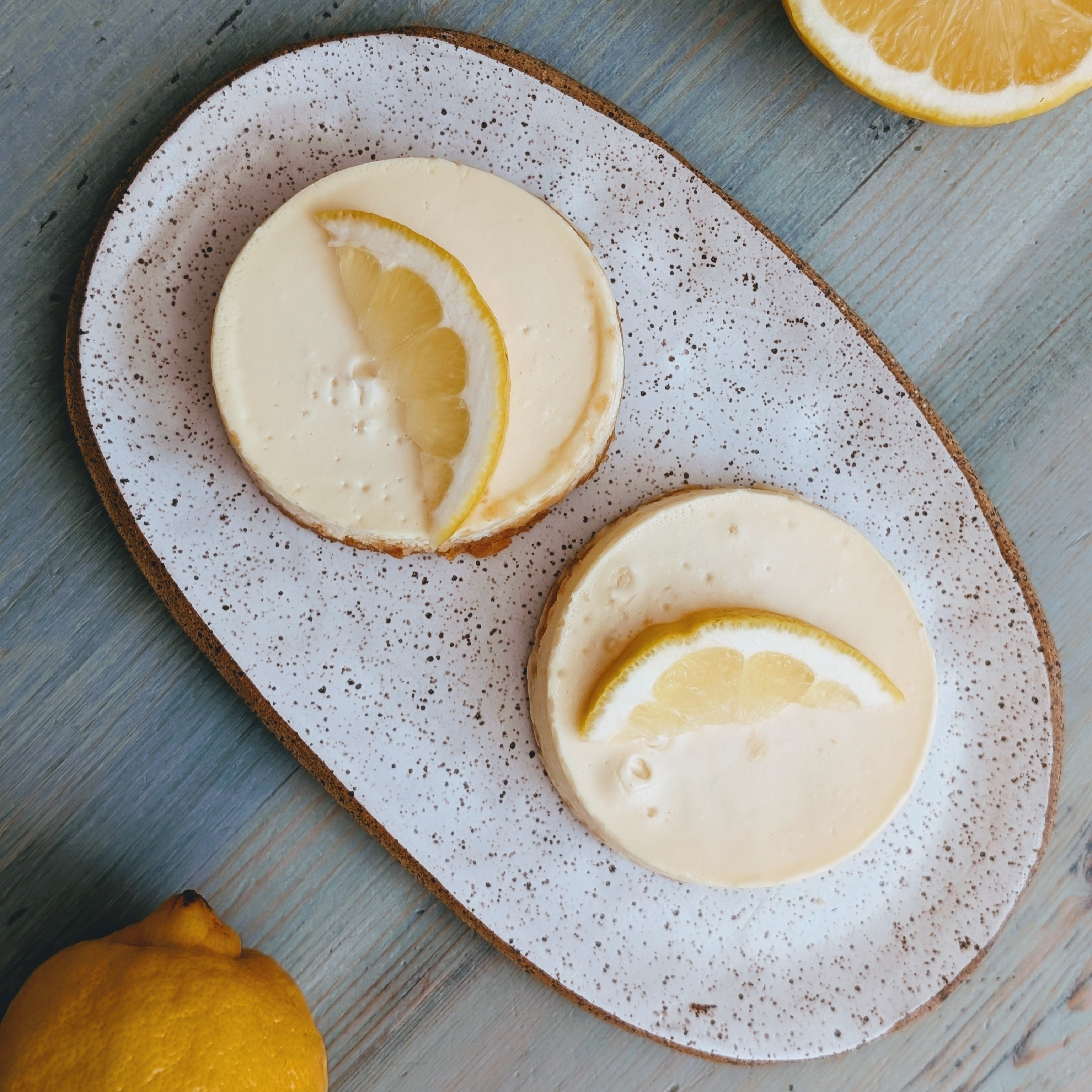 Two mini lemon cheesecakes on a speckled oval plate, each topped with a lemon wedge; whole lemon and lemon half nearby.