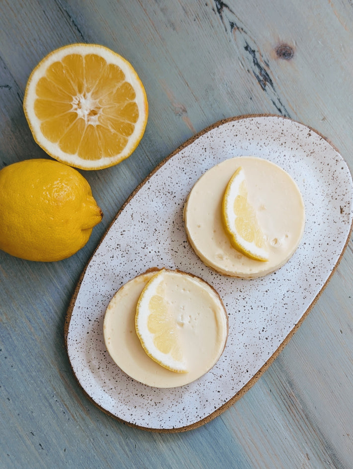 Two lemon slices on Wildgrain Lemon Cheesecakes with lemons on the side on a speckled ceramic plate.