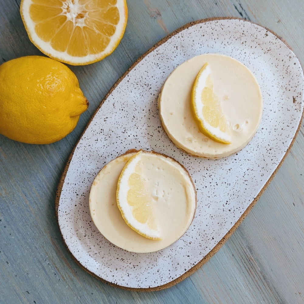 Two mini lemon cheesecakes on a speckled oval plate with lemon halves and a whole lemon on a blue wooden surface.