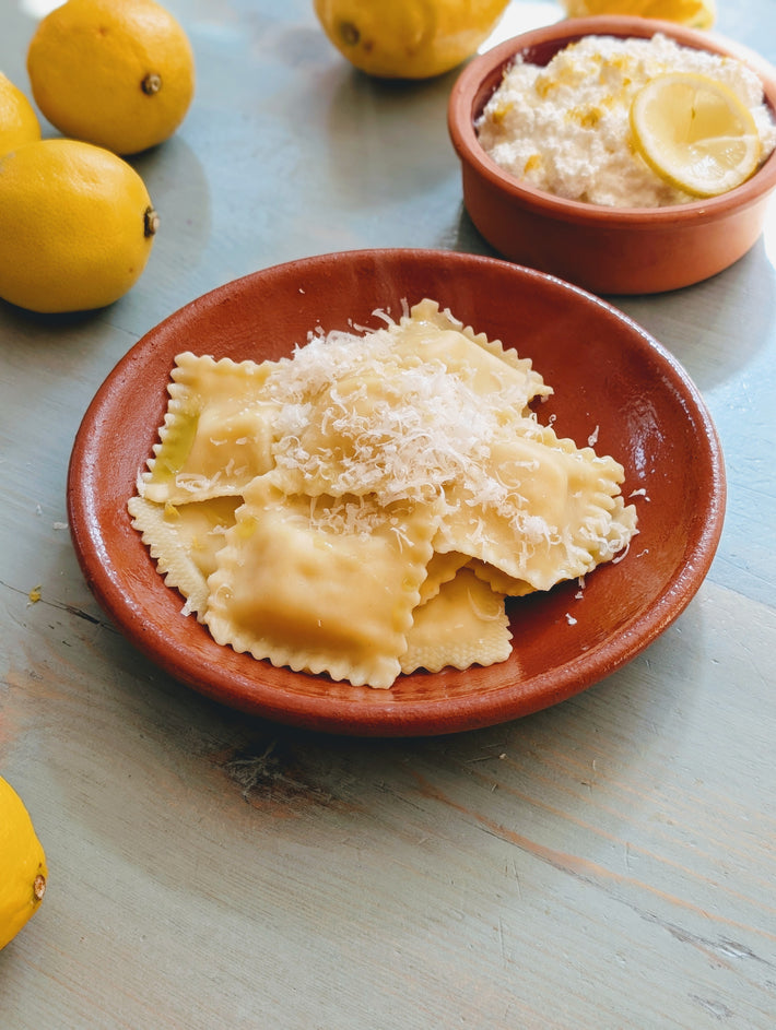 Ricotta and lemon ravioli on a plate with grated cheese, fresh lemons, and a bowl of ricotta.