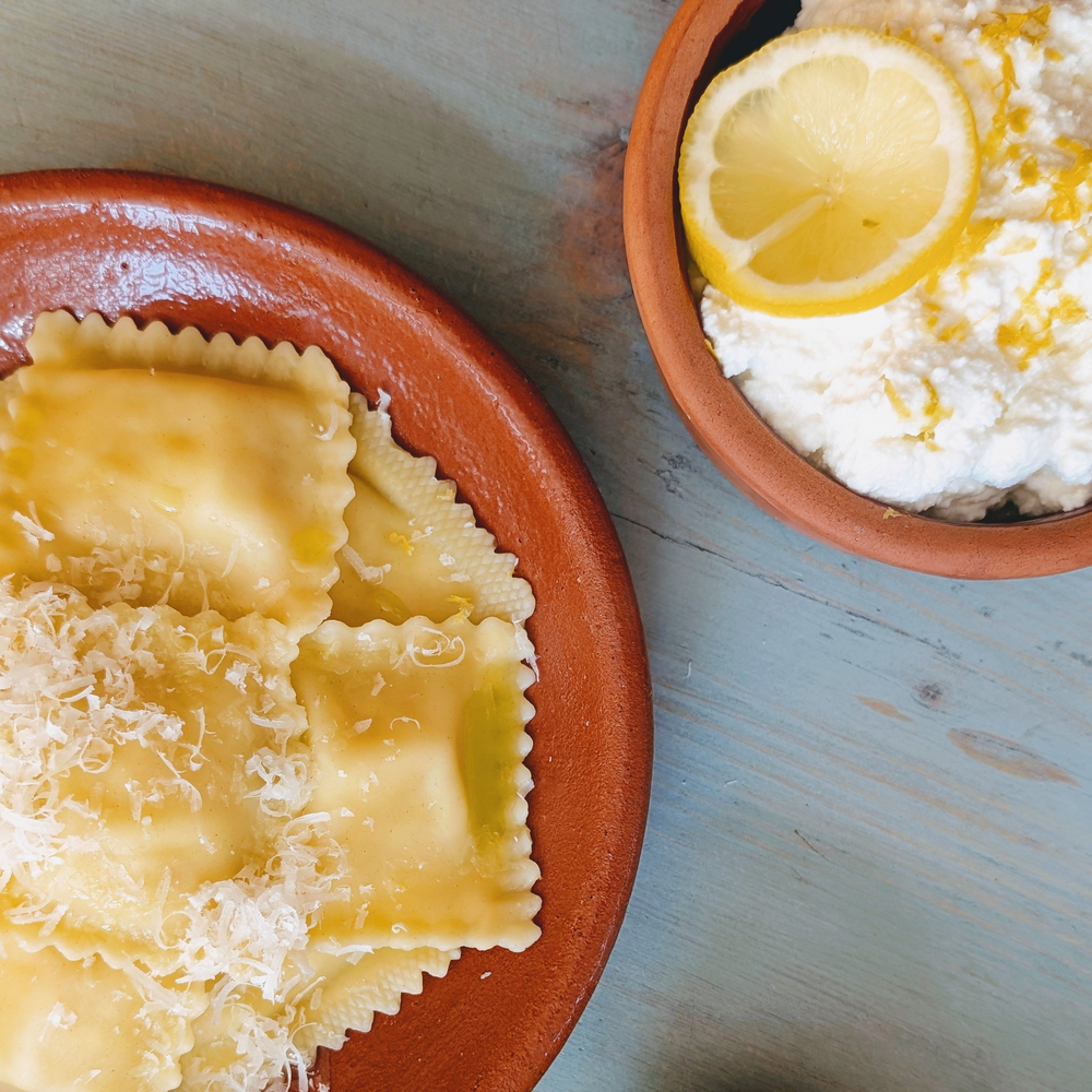 Buttery ravioli with grated Parmesan on a terracotta plate beside a bowl of ricotta topped with lemon slices