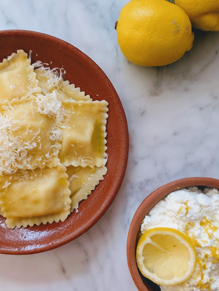 Plate of ricotta and lemon ravioli topped with grated cheese, bowl of ricotta cheese with a lemon slice, and two whole lemons on a marble surface.