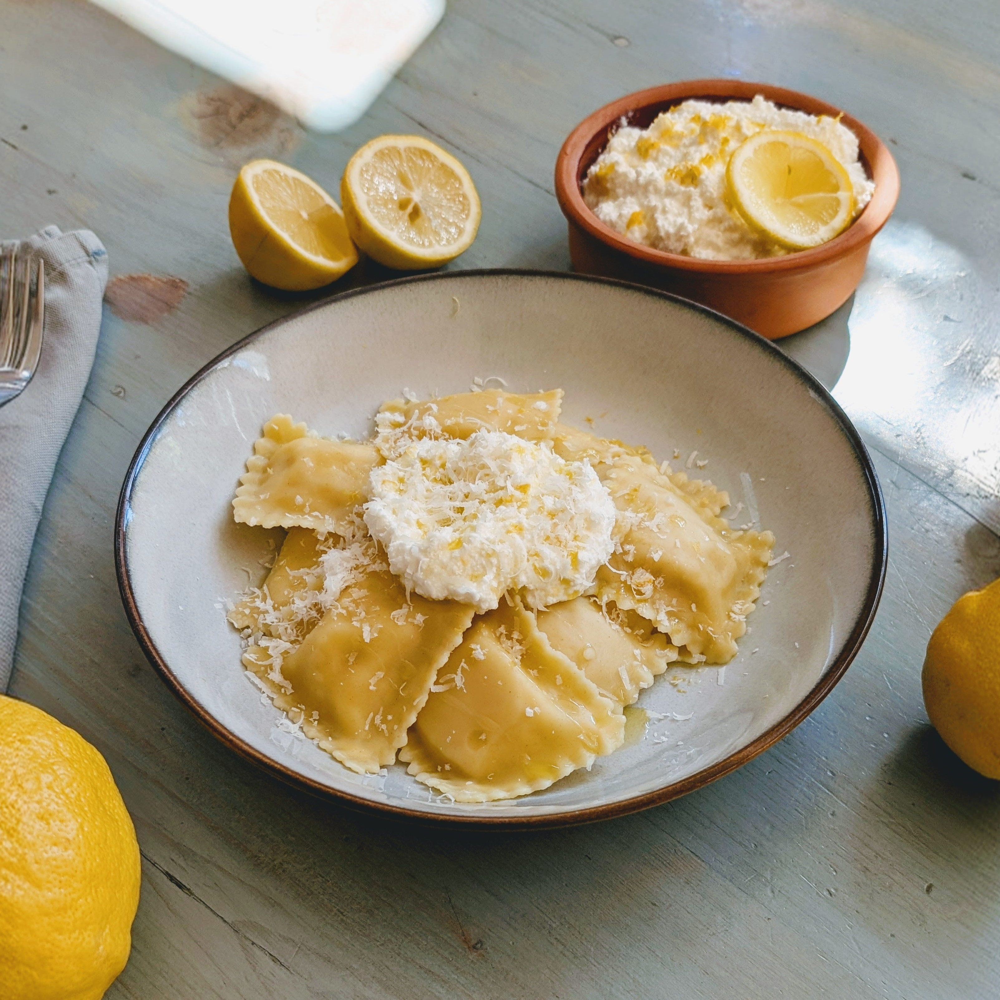 Cheese ravioli on a plate topped with ricotta and lemon zest, with a bowl of ricotta and halved lemons on a light blue table.