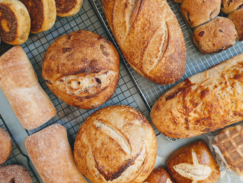 Assorted artisanal breads — round sourdough loaves, baguette-style loaves and rolls cooling on wire racks.