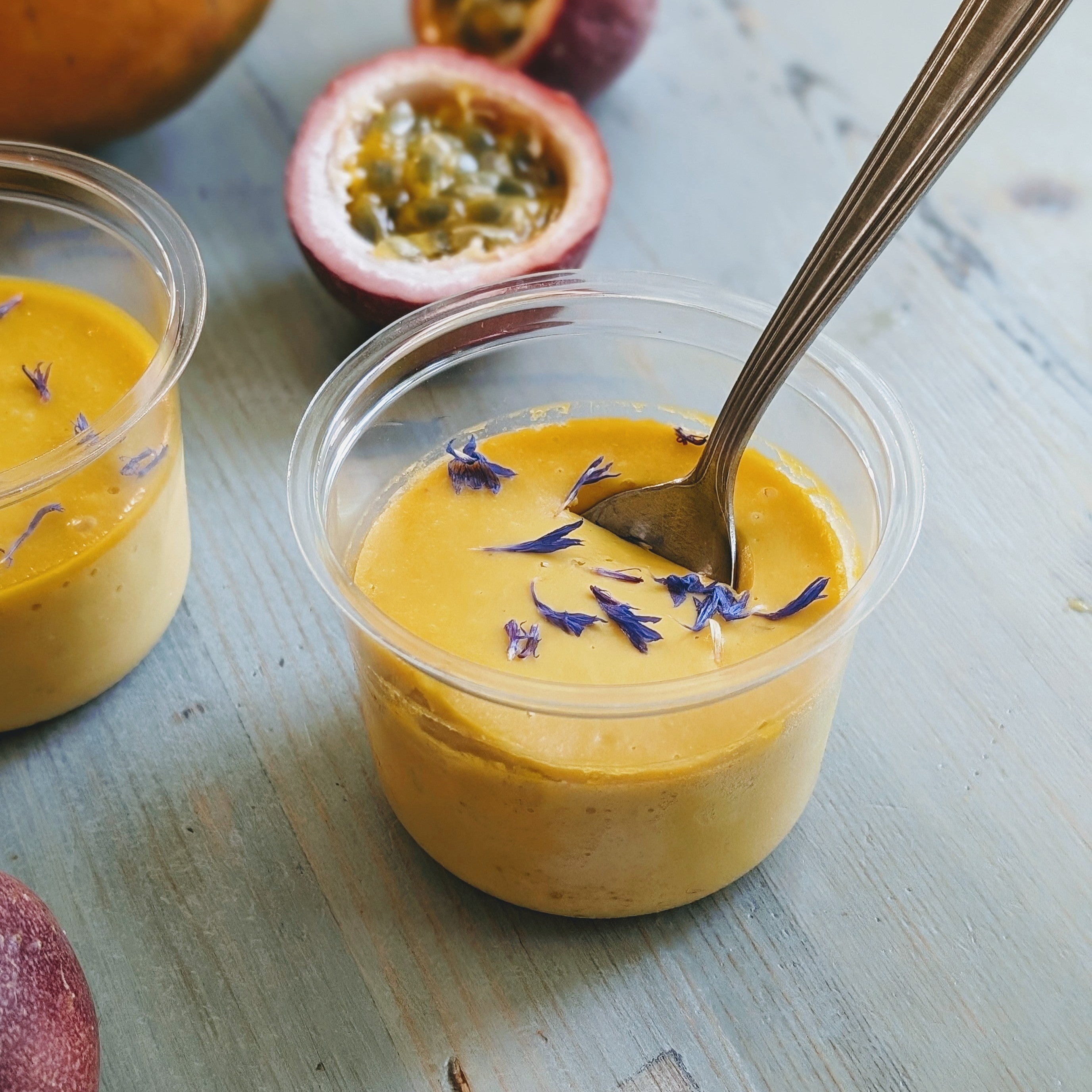 Plastic cup of passionfruit mousse topped with blue cornflower petals and a spoon; halved passionfruit in background