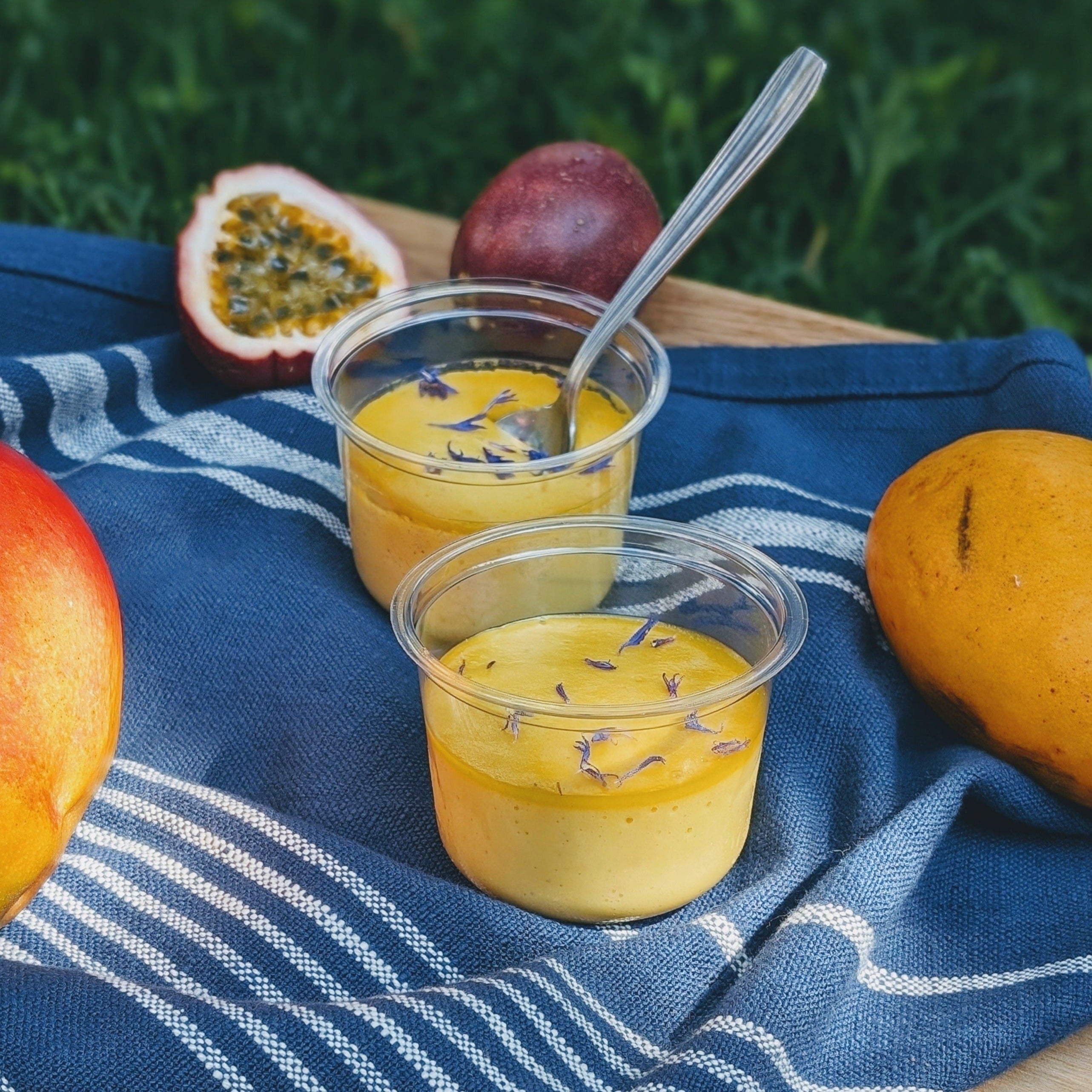 Two cups of mango and passion fruit mousse garnished with cornflower petals, surrounded by mangoes and passion fruits on a blue cloth.