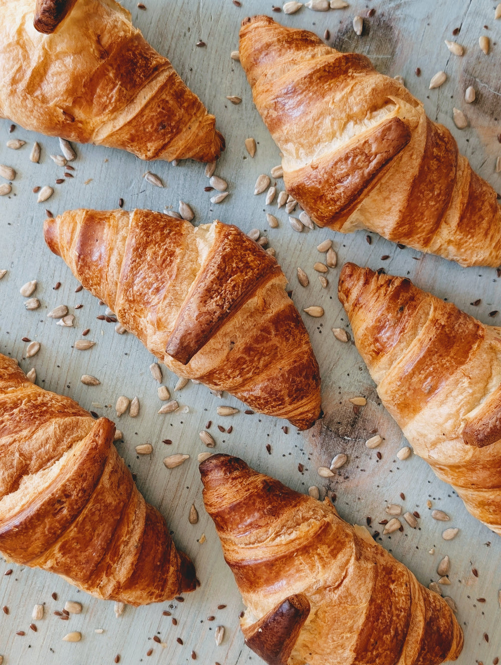 Freshly baked golden croissants scattered on a pale wooden board with sunflower seeds sprinkled around.