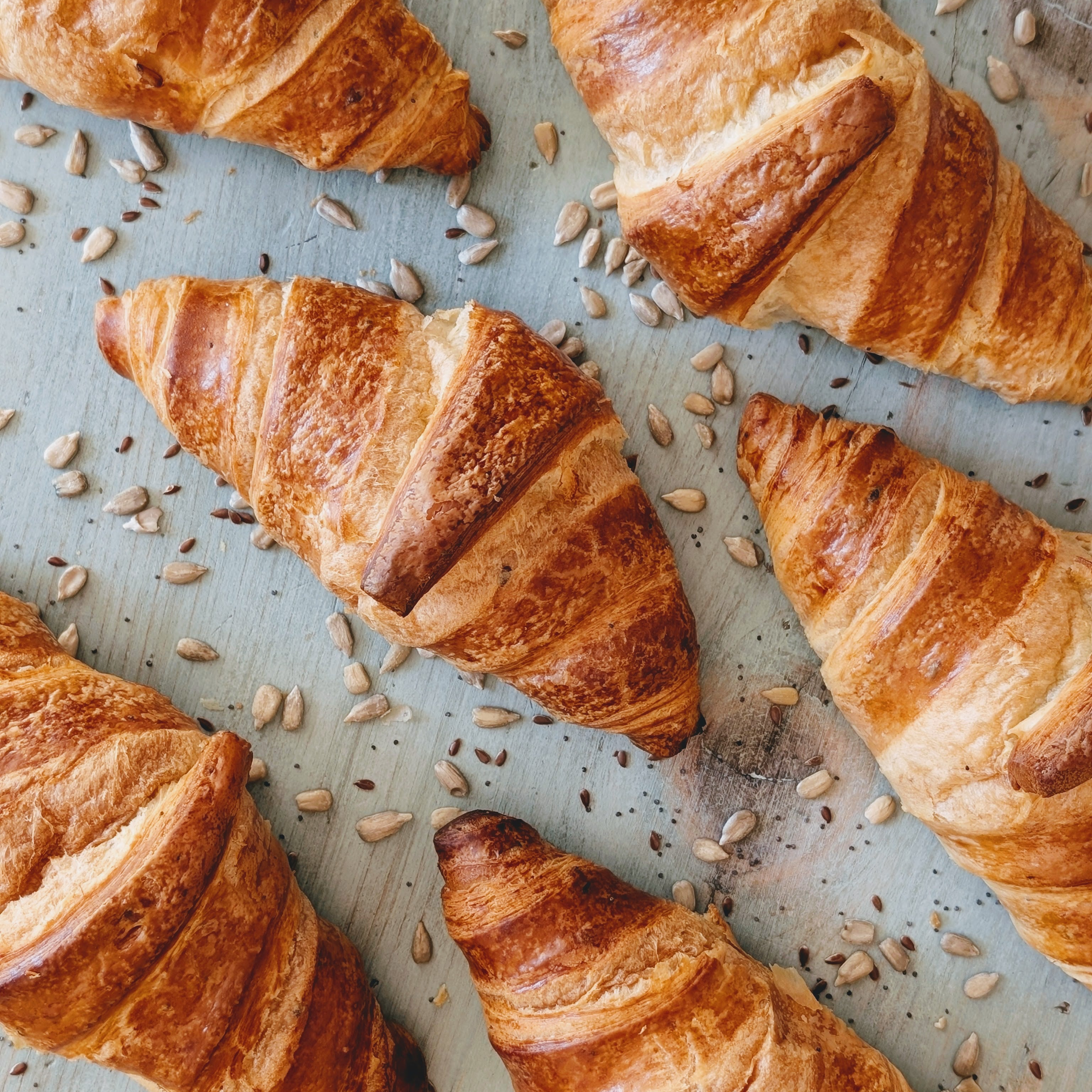 Six golden croissants on a light blue wooden surface sprinkled with sunflower seeds