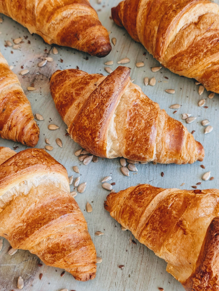 Golden-brown croissants on a pale wooden surface scattered with sunflower seeds.