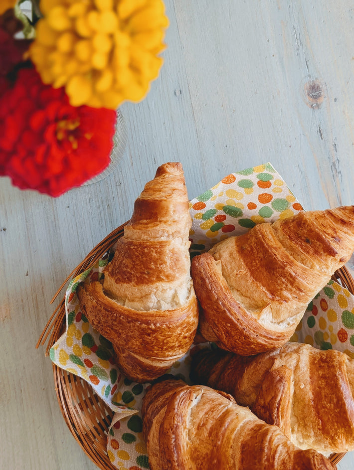 Four golden croissants in a wicker basket lined with a polka-dot napkin on a pale wooden table, blurred flowers at top left.