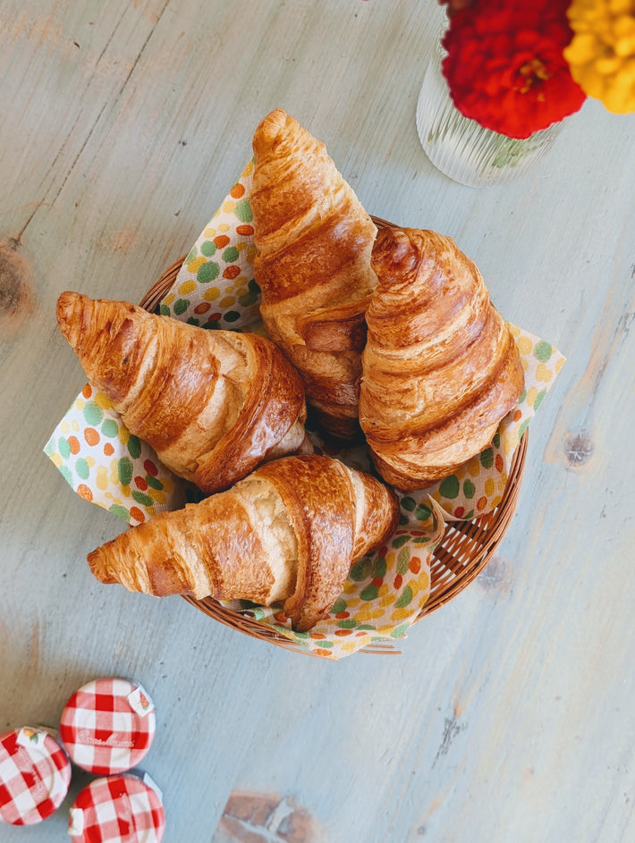 Four golden croissants in a wicker basket on a polka-dot napkin, jam jars with 'Bonne Maman' lids and a vase of flowers.