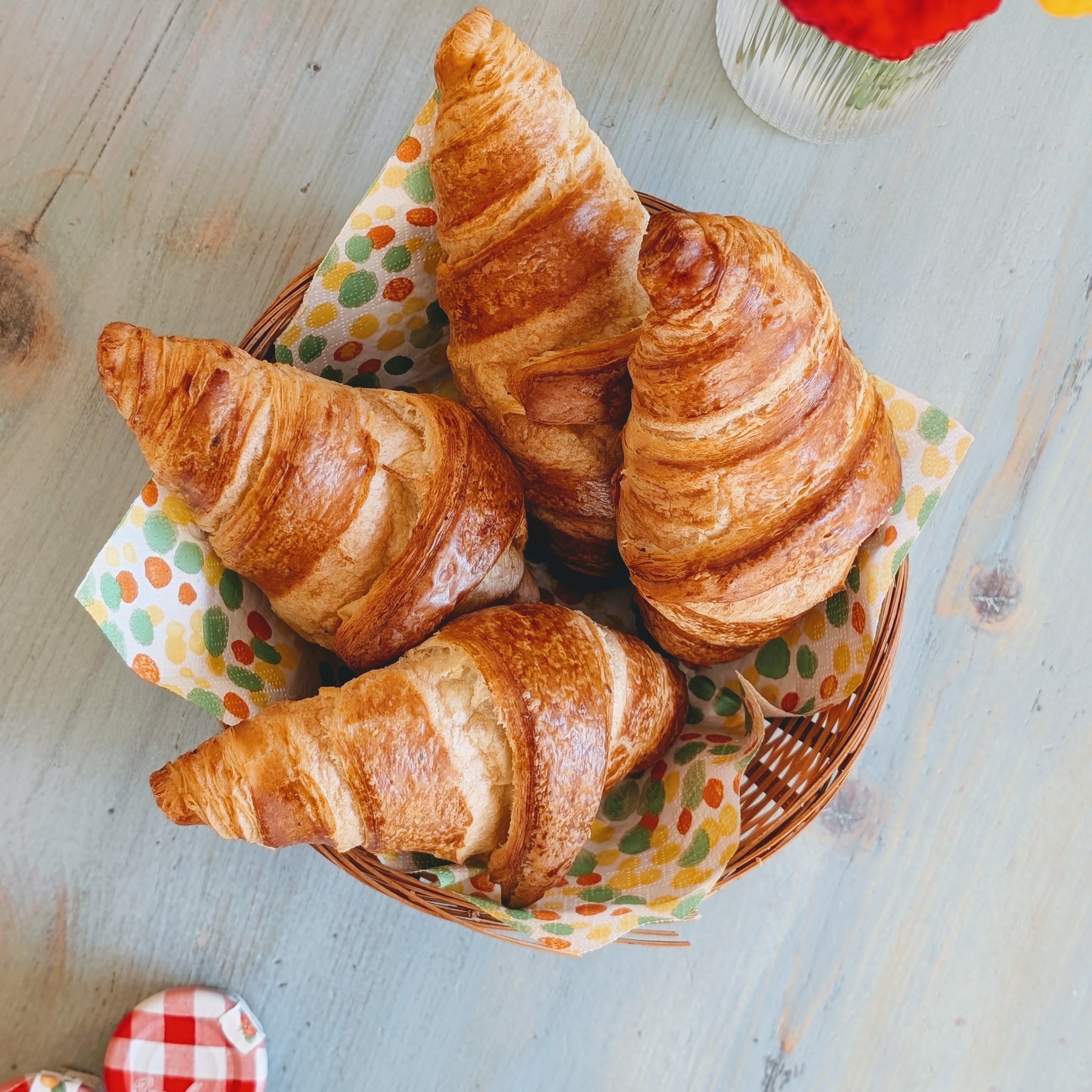 Four golden croissants in a wicker basket on a colorful polka-dot napkin on a pale blue wooden table.