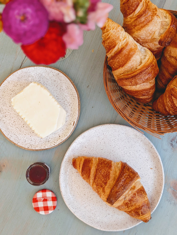 Overhead view of croissants on speckled plates, butter, jam jar with gingham lid reading 'Bonne Maman' and flowers.