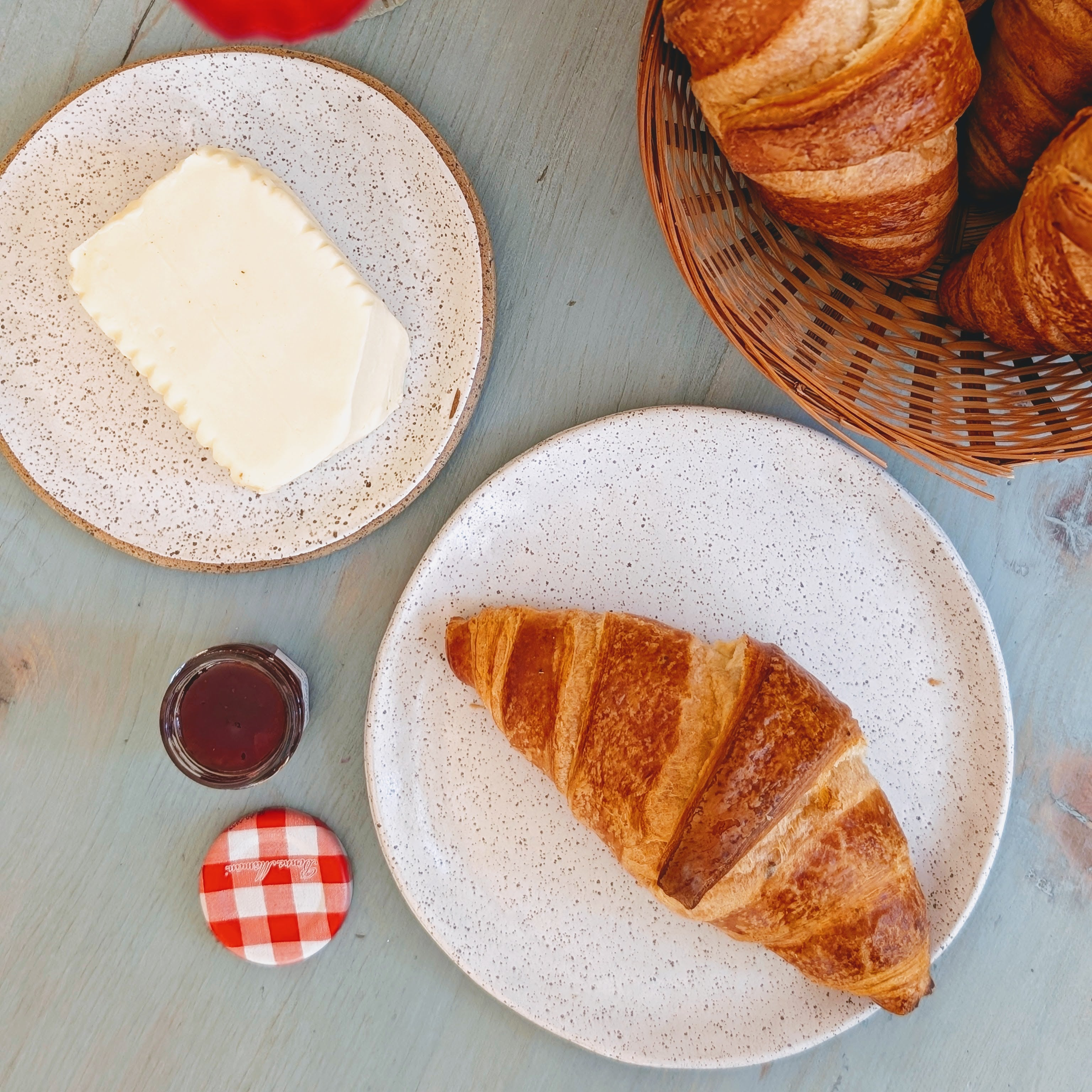 Croissant on speckled plate, butter on a plate, jam jar with Bonne Maman gingham lid and a basket of croissants.