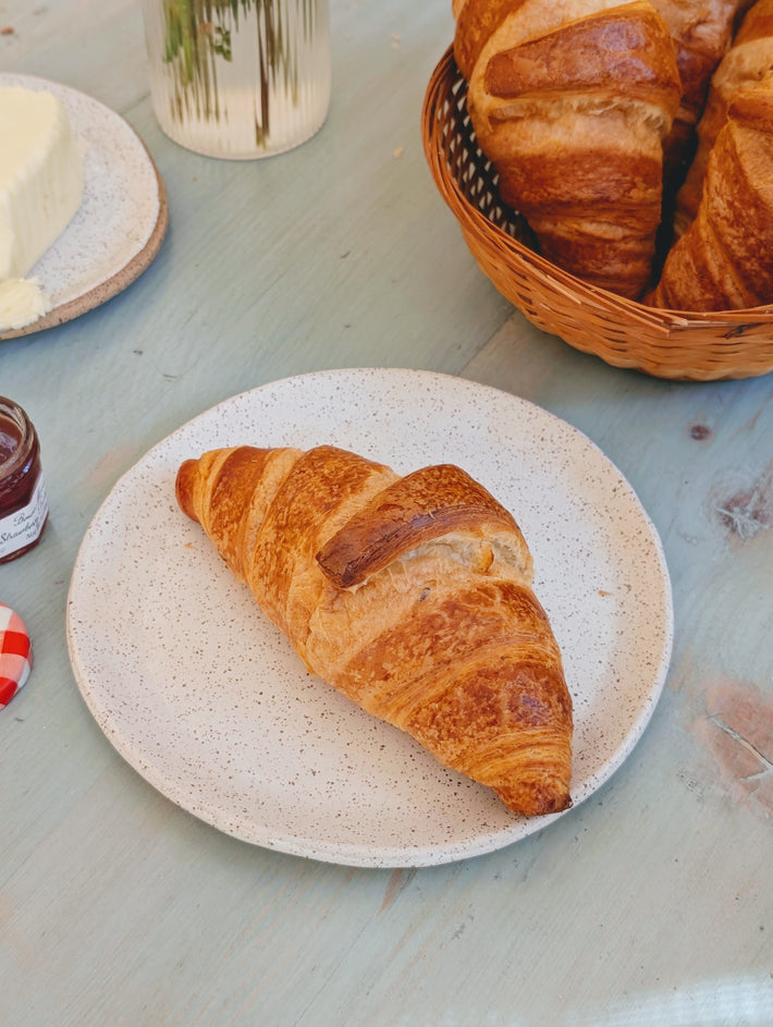 Buttery croissant on a speckled white plate, with basket of croissants and a butter dish on a light-blue wooden table.
