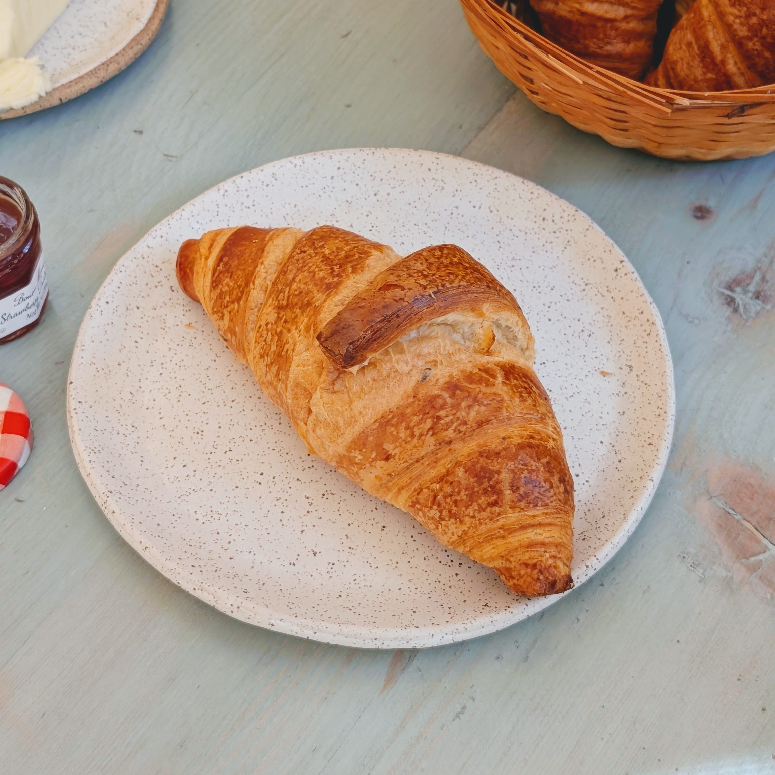 Buttery croissant on a speckled white plate on pale blue wooden table, jam jar with red-gingham lid and basket of croissants.