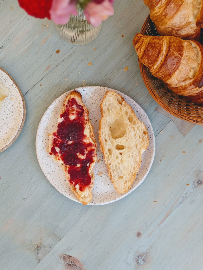 Two rustic bread slices on a plate, one spread with butter and red jam, the other plain; croissants in a basket nearby.