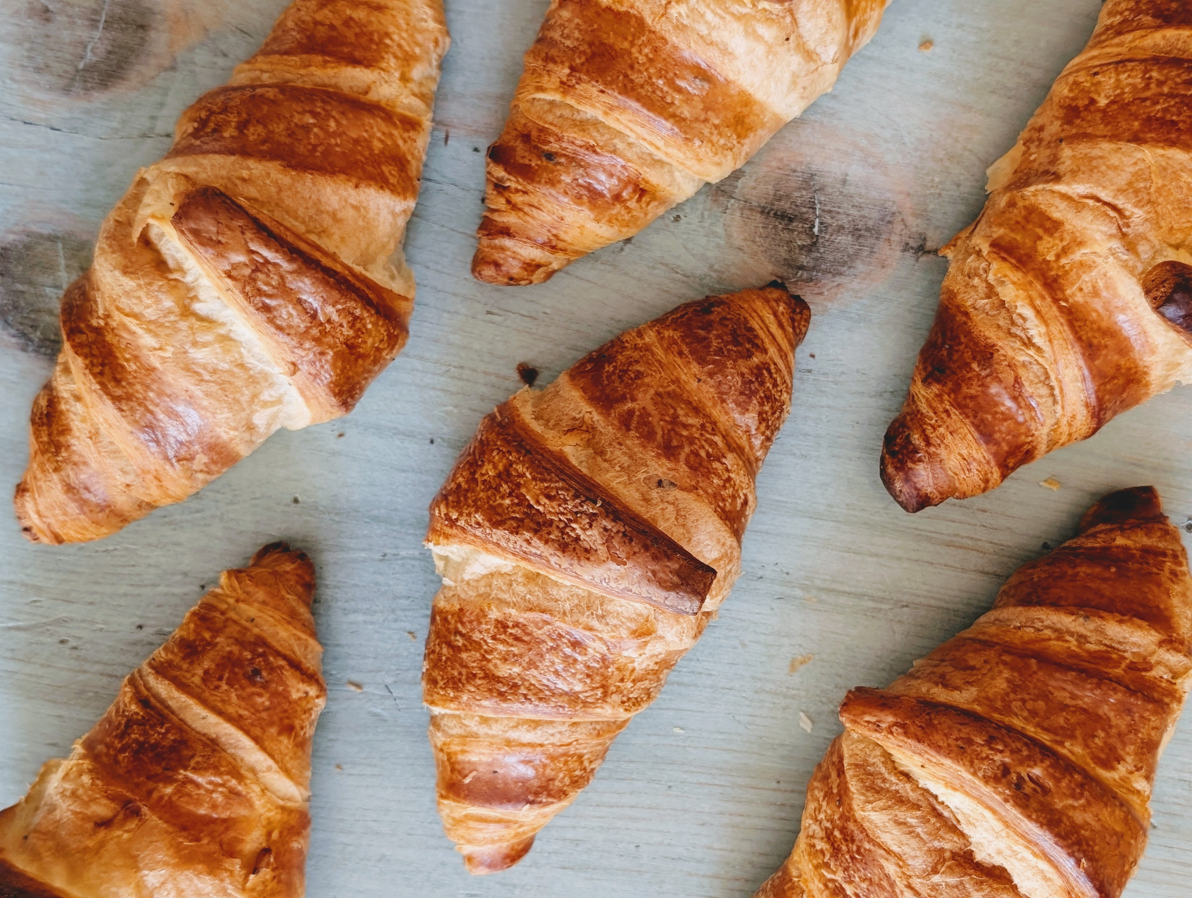 Six golden-brown croissants, whole and cropped, arranged on a pale wooden board.