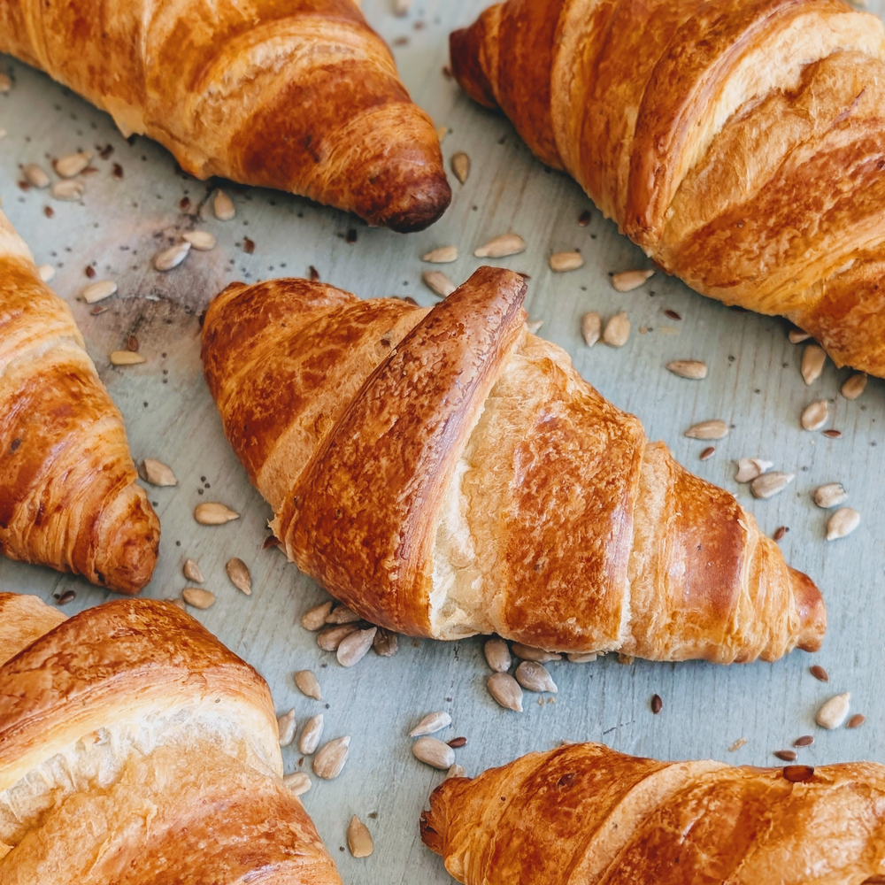 Buttery golden croissants on a pale blue surface sprinkled with sunflower seeds.