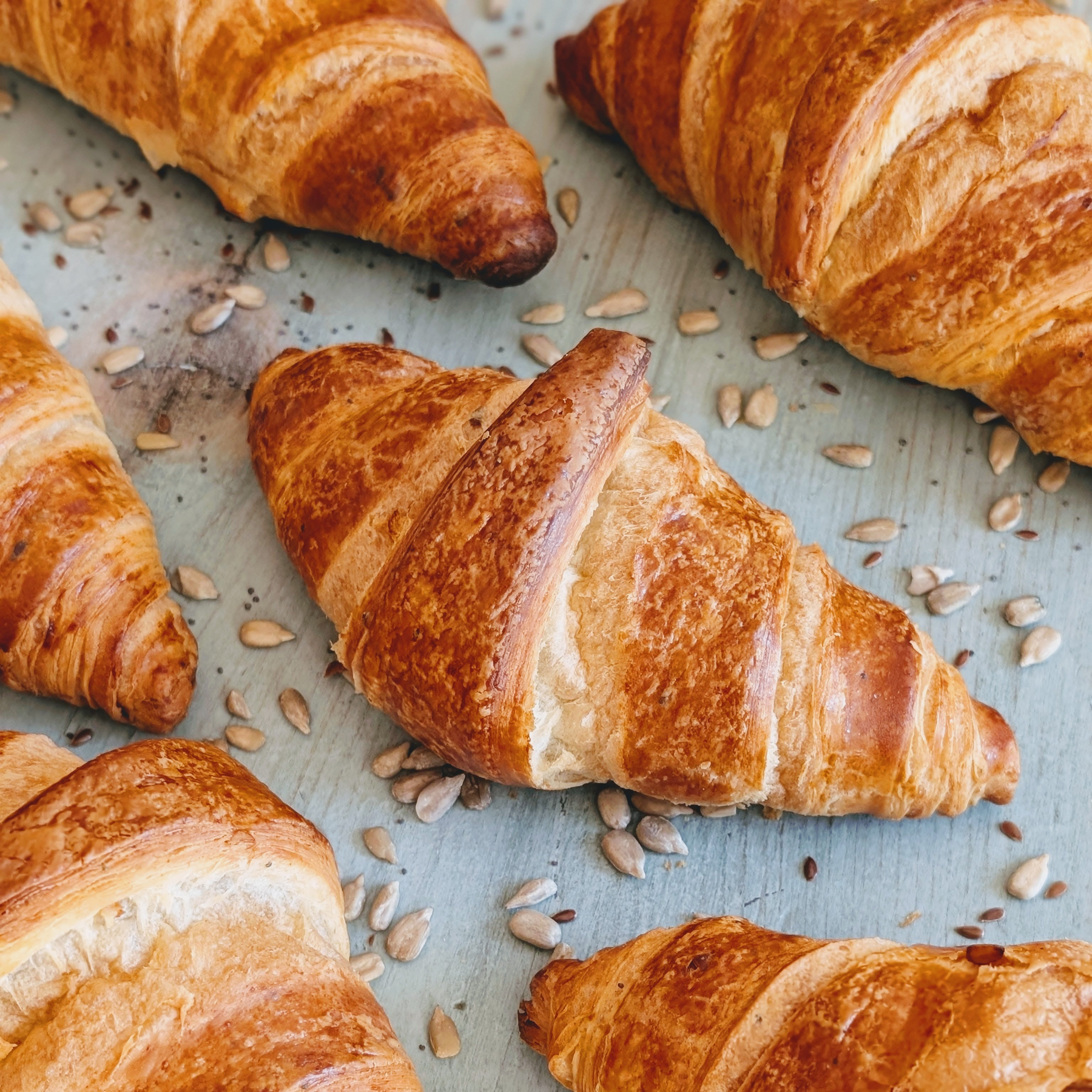 Buttery golden croissants on a pale blue surface sprinkled with sunflower seeds.