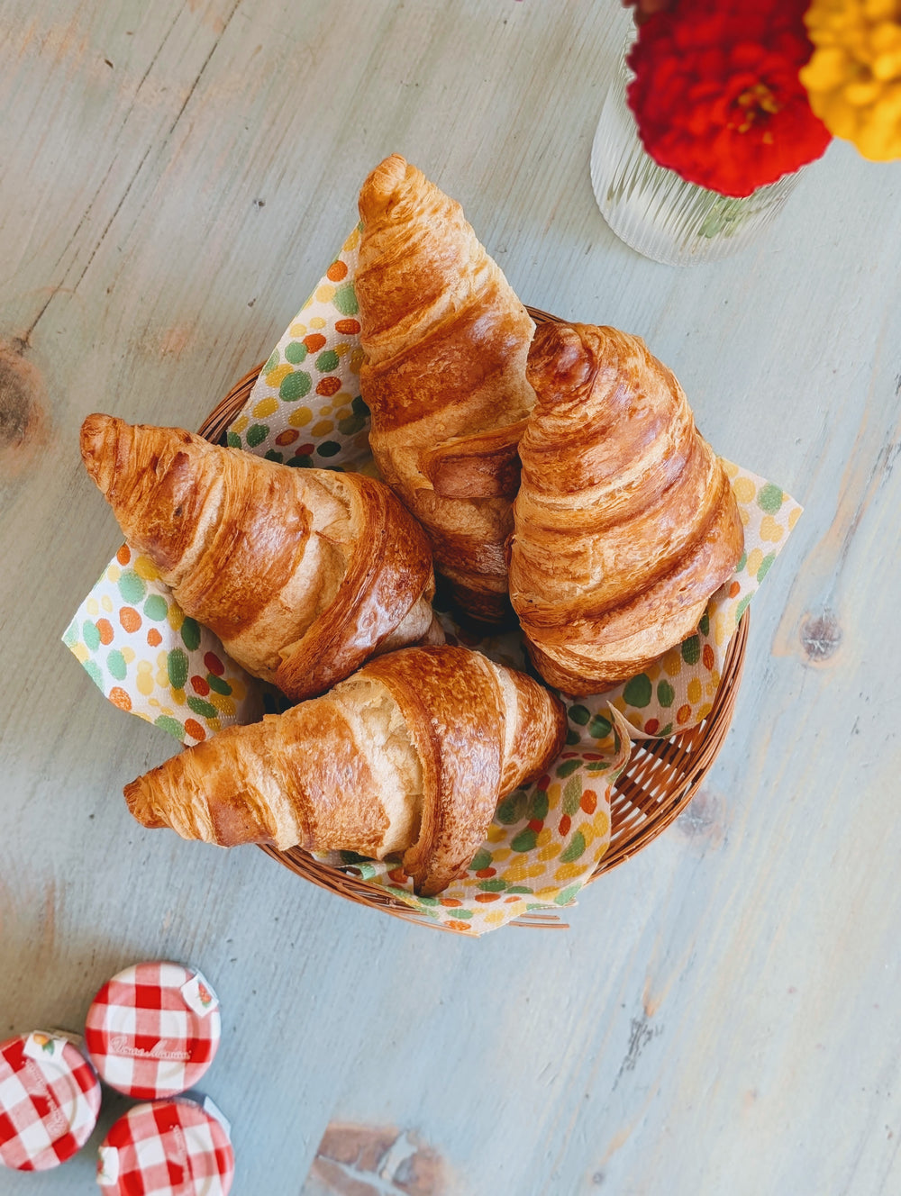 Four golden croissants in a wicker basket on a polka-dot napkin; three jam jar lids at lower left labeled 'Bonne Maman'.