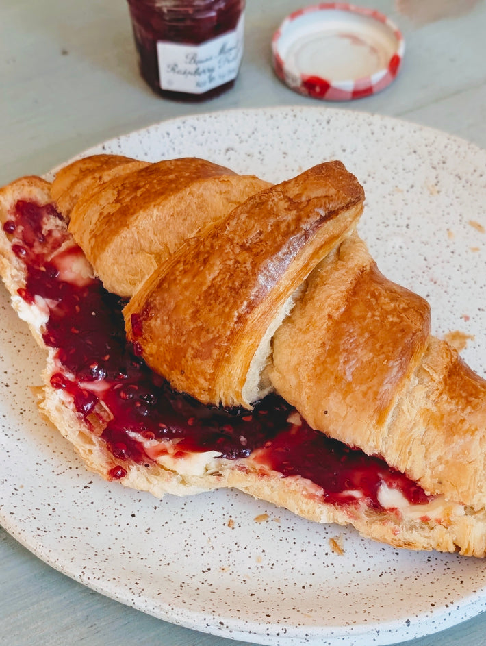 Halved croissant spread with butter and raspberry jam on a speckled plate, jam jar and lid blurred in background.