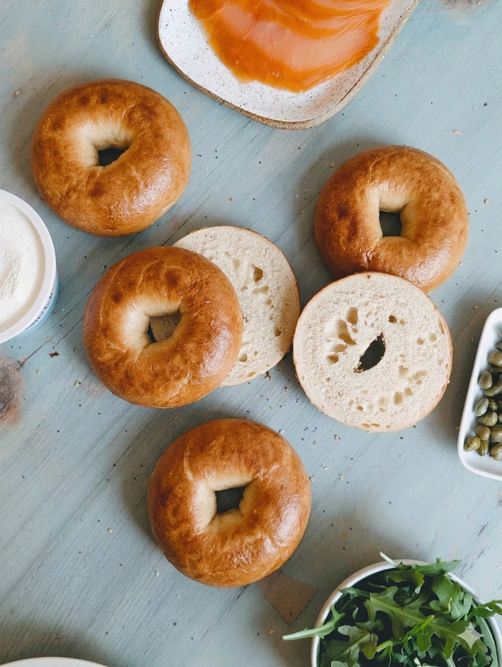 Overhead view of four plain New York bagels, one sliced to show interior, on a blue tabletop with smoked salmon and capers