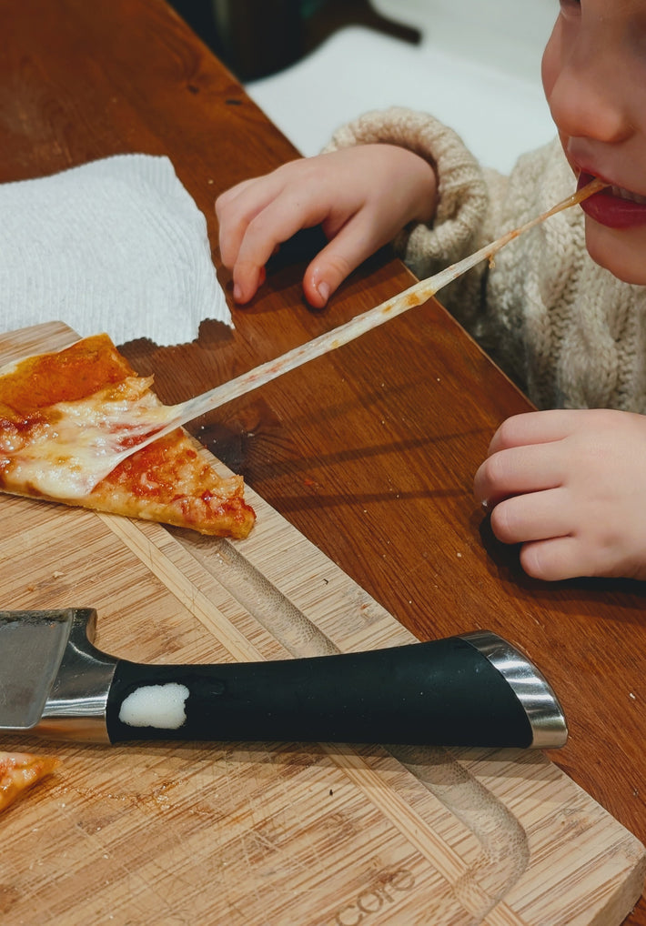 A child wearing a cozy sweater pulls a slice of pizza with cheese stretching, with a pizza cutter on the side.