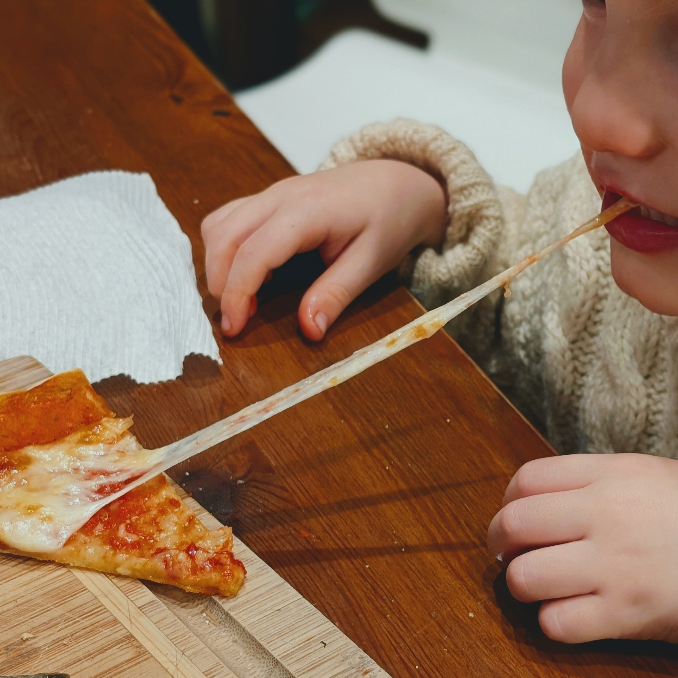 Child stretching a long strand of melted cheese from a pizza slice on a wooden table, hands and napkin visible.