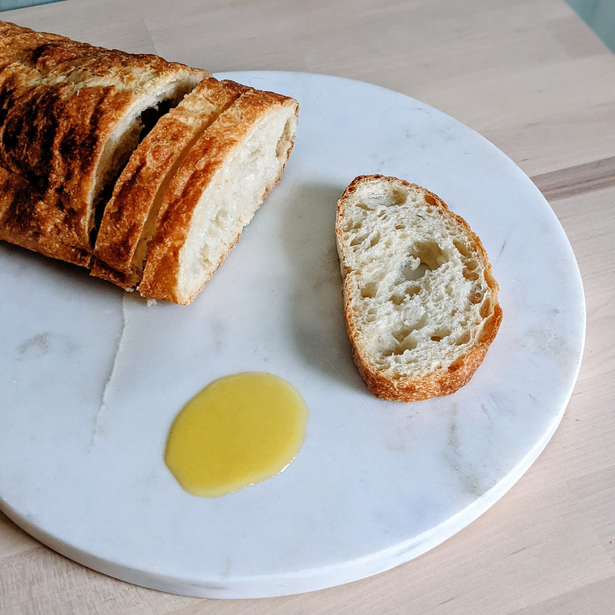 Sliced crusty baguette and one slice on a round marble board with a small pool of olive oil.