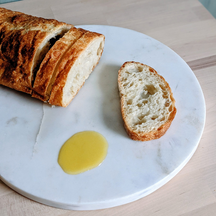 Sliced crusty baguette and one slice on a round marble board with a small pool of olive oil.
